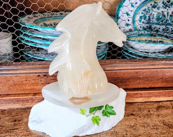 May include: A translucent, carved stone sculpture of a horse's head and neck. The sculpture sits on a white, rough-hewn base with green ivy accents. The horse is a light beige color, and the base has a small, oval-shaped brown accent.