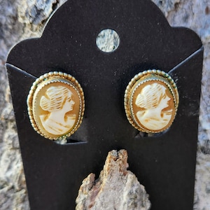 May include: A pair of gold tone earrings with a cameo design featuring a woman's profile in white.