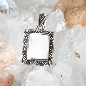 May include: A silver pendant featuring a rectangular, white mother-of-pearl centre. The pendant has a decorative border with small, dark accents. The bail is also silver and has a decorative design. The pendant is displayed on a surface of clear and tan crystals.