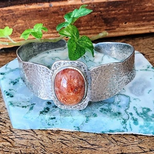 May include: A silver hammered cuff bracelet with a large oval-shaped orange sunstone cabochon.