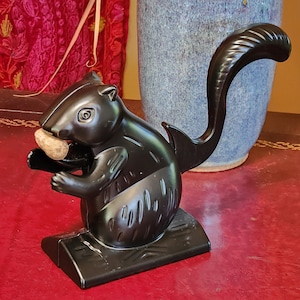 May include: A black metal squirrel nutcracker with a detailed design. The squirrel is holding a nut in its mouth and has a long tail.