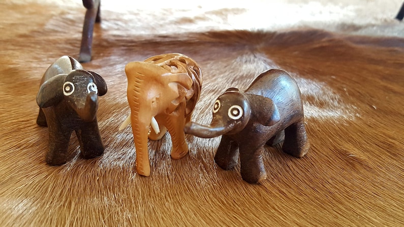 May include: Three wooden elephant figurines, one brown, one light brown, and one dark brown. The figurines are standing on a brown and white cowhide rug.