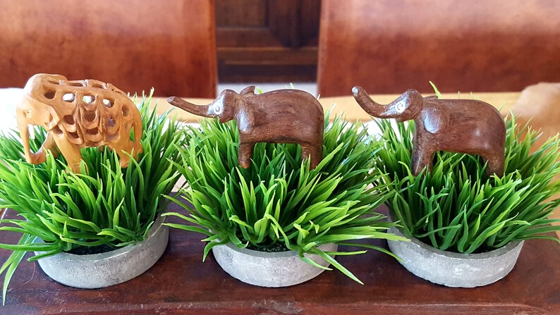 May include: Three wooden elephant figurines, each standing on a bed of green artificial grass in a gray concrete pot. The elephants are carved in different styles, with one being a more intricate design.