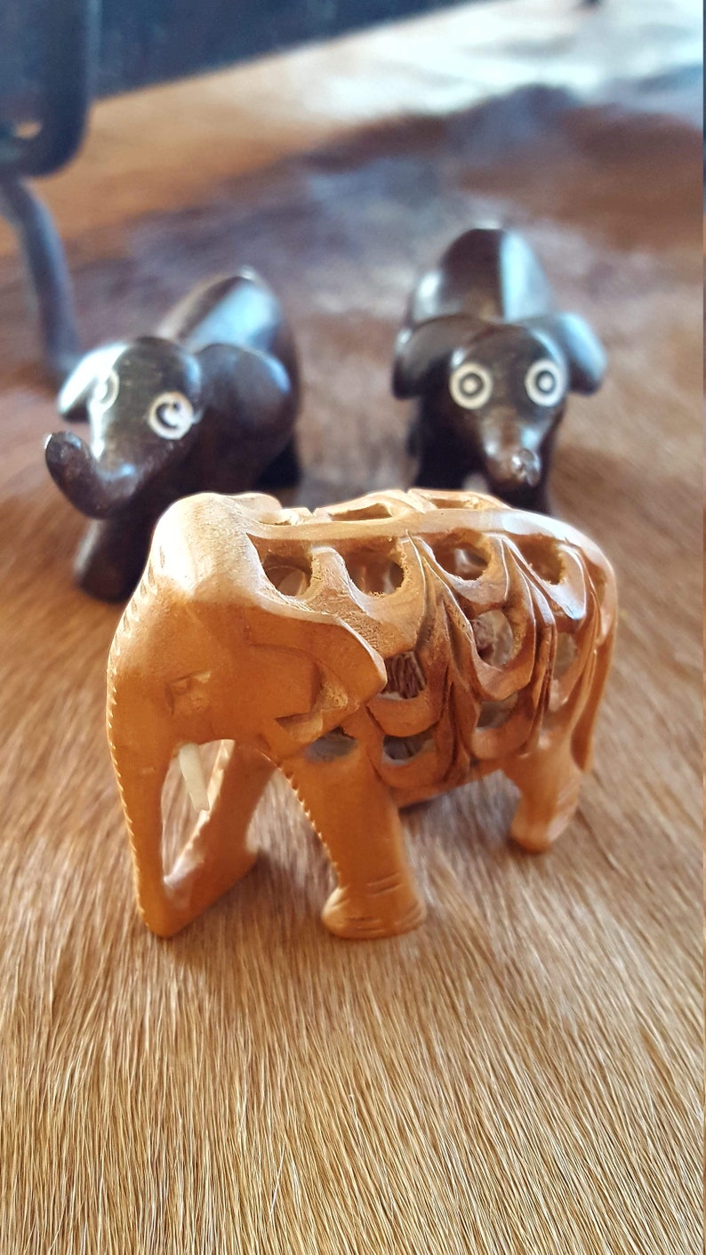 May include: Three wooden elephant figurines. Two are dark brown and one is light brown with intricate carvings. The light brown elephant is in focus and the other two are out of focus.