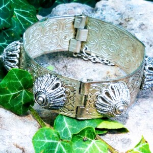 May include: A silver bracelet with a hinged clasp and decorative floral patterns. The bracelet has four decorative accents on the front, each with a raised, circular design.