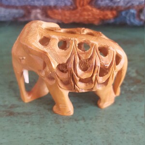 May include: A small, intricately carved wooden elephant figurine. The elephant is light brown in color and has a detailed design on its back. The figurine is standing on a green surface.