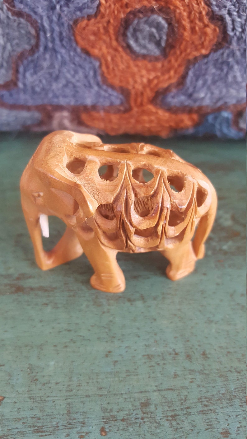 May include: A small, intricately carved wooden elephant figurine. The elephant is light brown in color and has a detailed design on its back. The figurine is standing on a green surface.