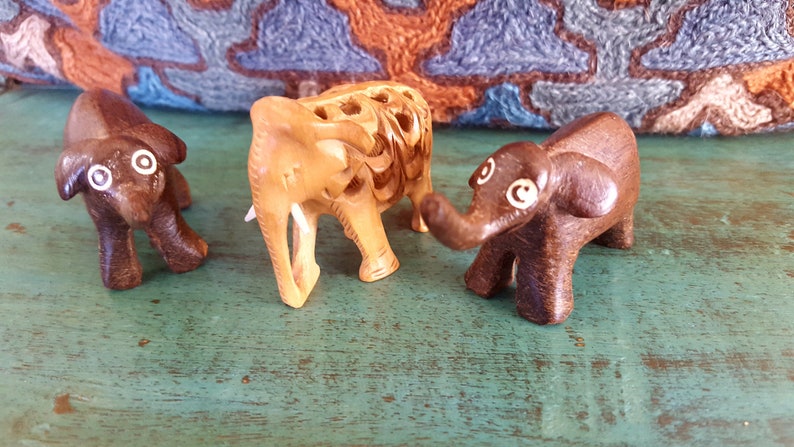 May include: Three wooden elephant figurines, one is light brown with intricate carvings, the other two are dark brown with white painted eyes.