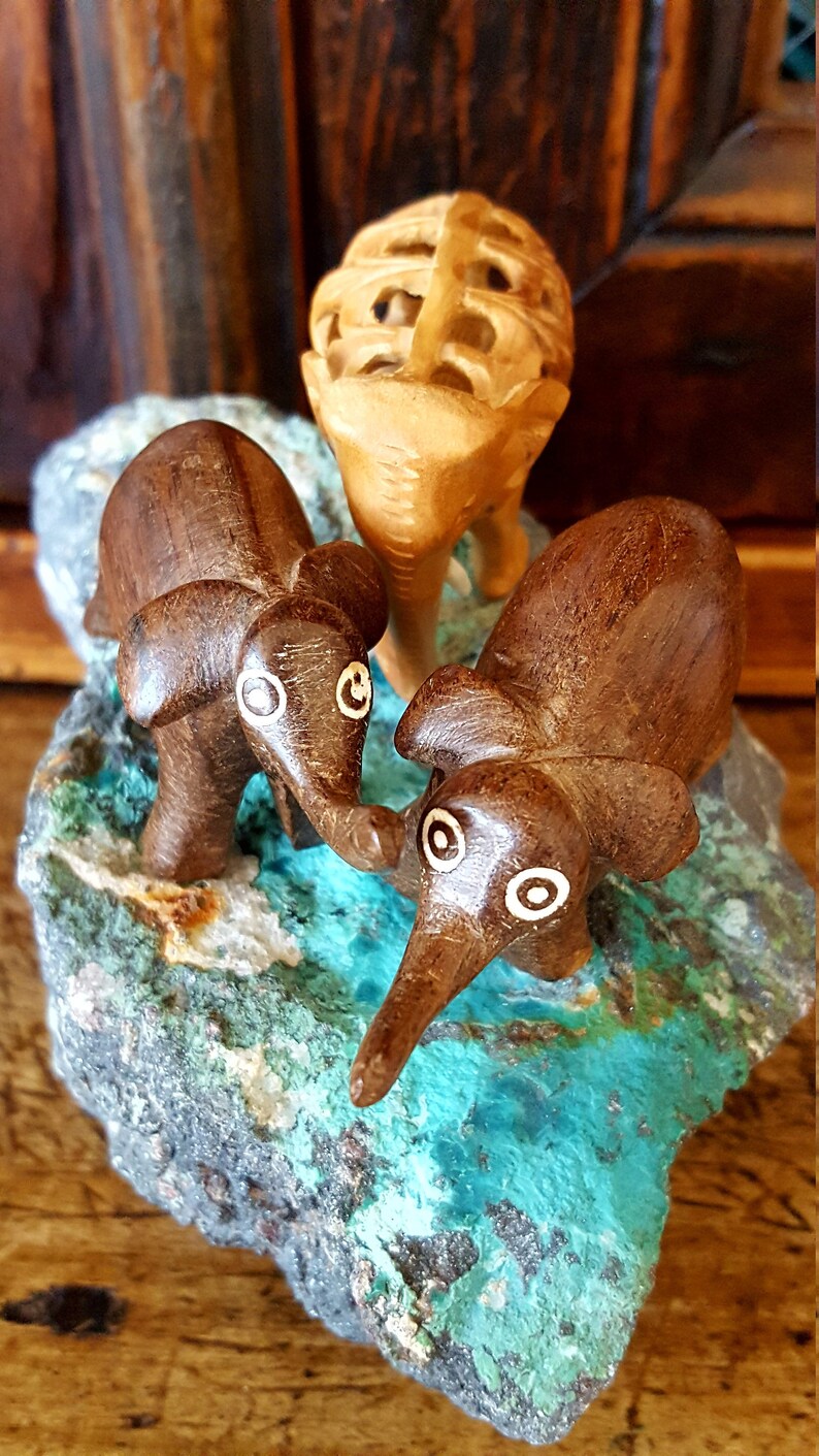 May include: Three wooden elephant figurines, two brown and one light brown, are arranged on a turquoise and gray rock. The elephants have white painted eyes and are carved in a simple style.