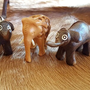 May include: Three wooden elephant figurines, one brown, one dark brown, and one light brown, with white accents around the eyes. The figurines are arranged on a brown and white fur surface.