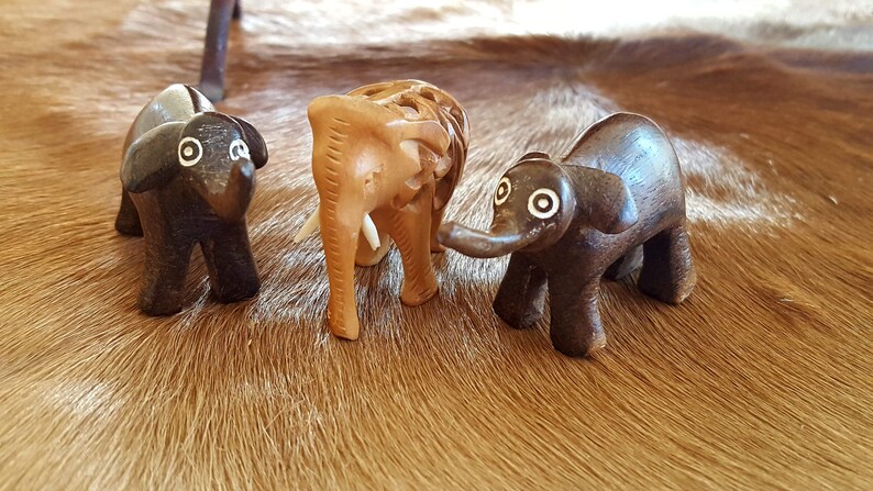 May include: Three wooden elephant figurines, one brown, one dark brown, and one light brown, with white accents around the eyes. The figurines are arranged on a brown and white fur surface.