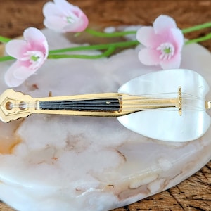 May include: A white and gold musical instrument brooch with a black inlay. The brooch is shaped like a violin and is set on a white and brown agate stone.
