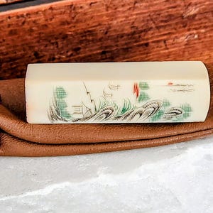 May include: A small, rectangular, ivory-colored stamp with a painted landscape scene of green trees, water, and red accents. The stamp is in a brown leather pouch with a purple drawstring.