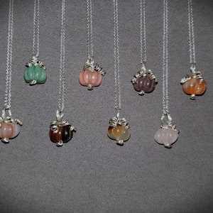 May include: A set of eight silver necklaces with gemstone pumpkin pendants. The pumpkins are in various colors, including green, pink, brown, and orange.