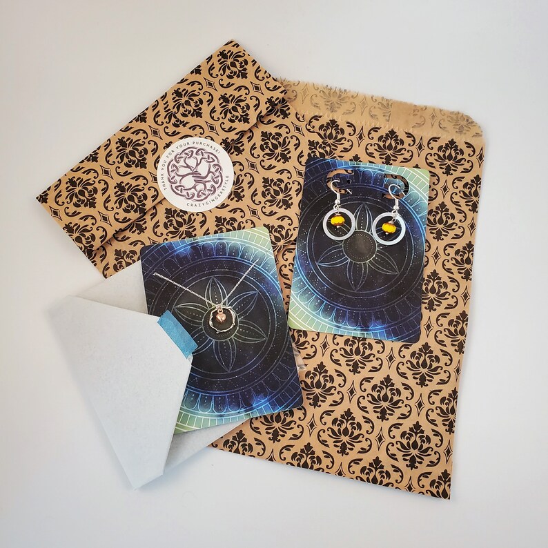 May include: A brown paper bag with a black and white patterned design. The bag contains two cards with a necklace and earrings. The cards have a blue and black background with a mandala design. The text on the bag reads "Thank you for your purchase, CraftyCorners".