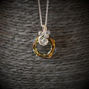 May include: A silver necklace with a pendant featuring a circular, faceted crystal in shades of gold and blue. The pendant is attached to a silver chain and has a decorative silver swirl design above the crystal.