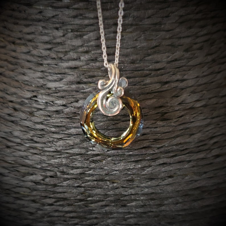May include: A silver necklace with a pendant featuring a circular, faceted crystal in shades of gold and blue. The pendant is attached to a silver chain and has a decorative silver swirl design above the crystal.