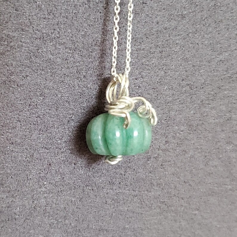 May include: A silver chain necklace with a green jade pumpkin pendant. The pumpkin is carved with ridges and has a silver wire stem.