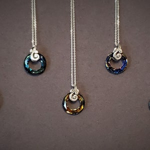 May include: A collection of silver necklaces with circular pendants. Each pendant features a ring of iridescent crystals in various colors, including blue, green, and gold. The necklaces hang against a dark brown background.