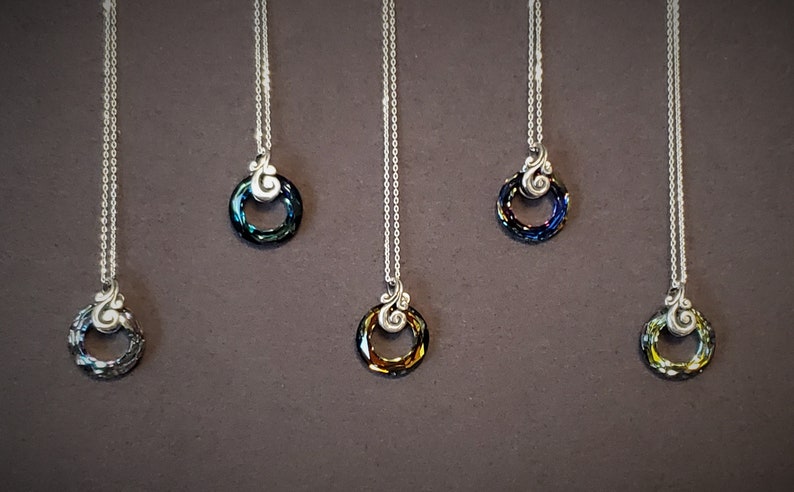 May include: A collection of silver necklaces with circular pendants. Each pendant features a ring of iridescent crystals in various colors, including blue, green, and gold. The necklaces hang against a dark brown background.
