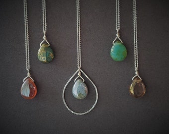 May include: Five silver necklaces with different colored gemstone pendants. The pendants are all teardrop shaped and have a metallic sheen. The necklaces are on a gray background.