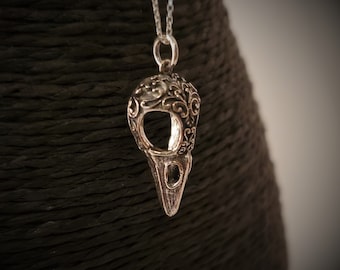 May include: A silver-colored pendant necklace featuring a raven's skull design. The pendant has intricate filigree detailing on the top and a pointed beak. The necklace hangs on a delicate silver chain, showcasing a gothic or fantasy theme.