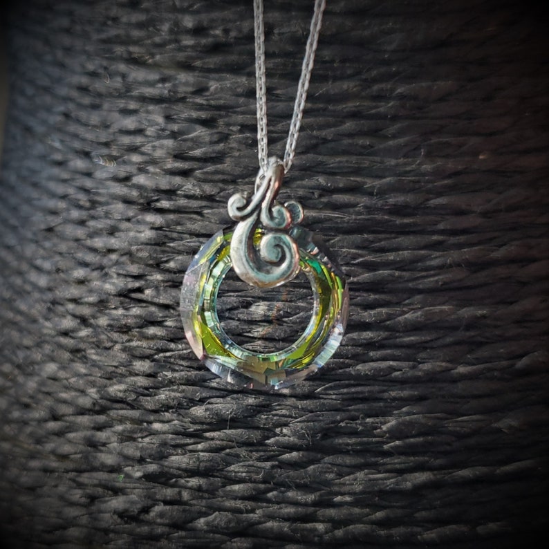 May include: A silver necklace featuring a circular pendant with a green and yellow iridescent center. The pendant has a decorative silver swirl design at the top and hangs from a delicate silver chain. The necklace is displayed against a dark, textured background.