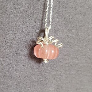 May include: A silver chain necklace with a pink glass pumpkin pendant. The pumpkin is detailed with silver wire and has a ribbed texture.