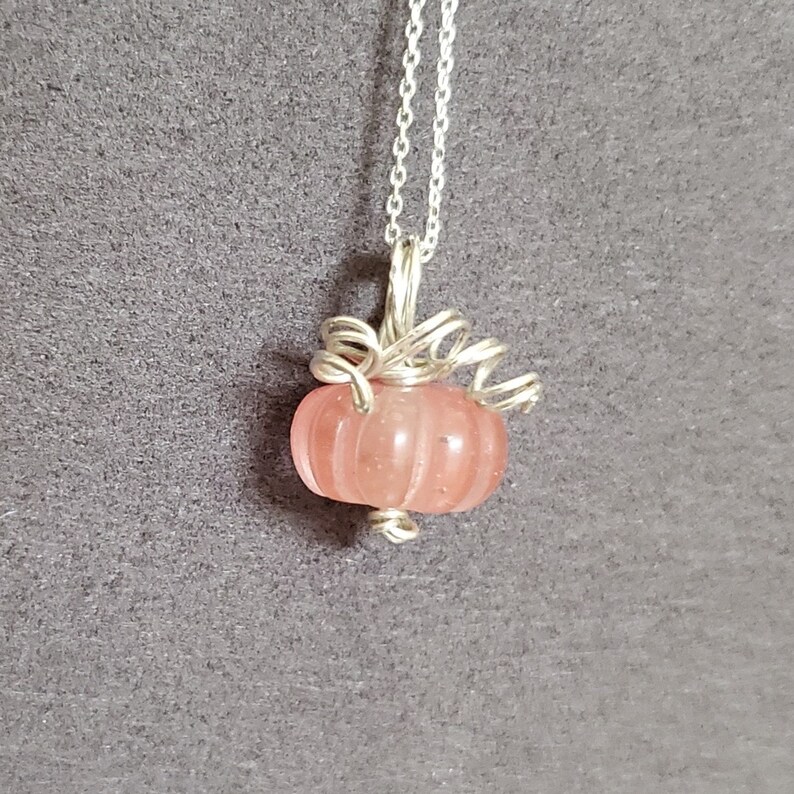 May include: A silver chain necklace with a pink glass pumpkin pendant. The pumpkin is detailed with silver wire and has a ribbed texture.