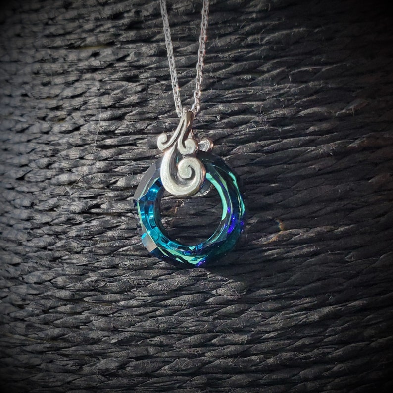 May include: A silver necklace with a pendant featuring a faceted, teal-colored crystal ring. The pendant has a decorative silver swirl design and hangs from a delicate chain. The background is a dark, textured surface.