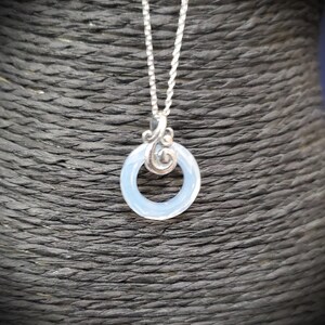 May include: A silver necklace featuring a pendant with a light blue, circular stone. The pendant has a decorative silver swirl design above the stone and hangs from a delicate silver chain. The necklace is displayed against a dark, textured background.