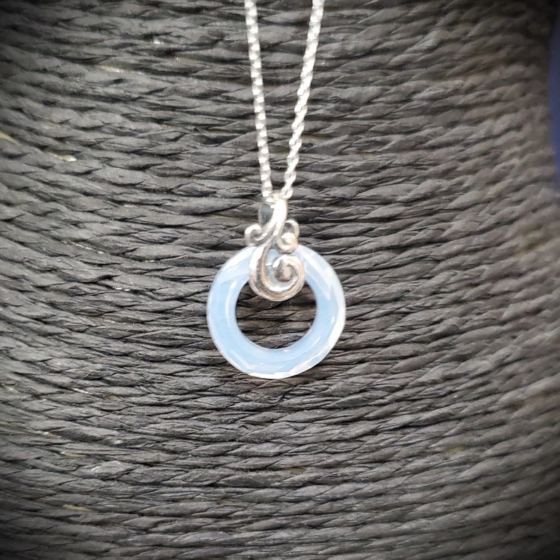 May include: A silver necklace featuring a pendant with a light blue, circular stone. The pendant has a decorative silver swirl design above the stone and hangs from a delicate silver chain. The necklace is displayed against a dark, textured background.