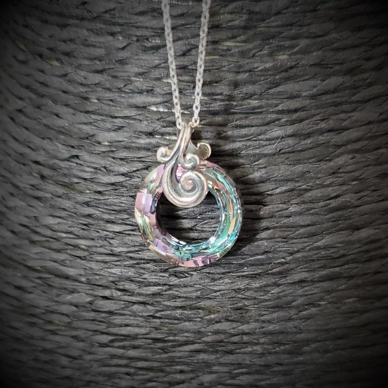 May include: A silver necklace featuring a circular pendant with iridescent crystals. The pendant has a decorative silver swirl design at the top. The chain is a delicate silver link. The crystals display shades of pink, blue, and green.