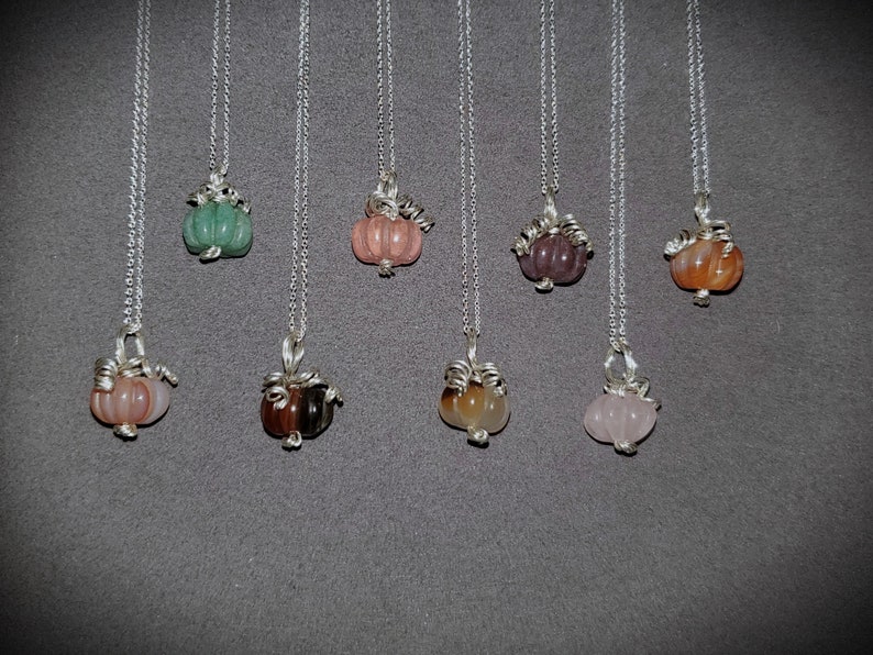May include: A set of eight silver necklaces with gemstone pumpkin pendants. The pumpkins are in various colors, including green, pink, brown, and orange.