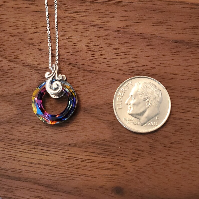 May include: A silver necklace with a circular pendant. The pendant is a ring of iridescent crystals, displaying a spectrum of colors. A U.S. dime is next to the necklace for size comparison. The necklace is on a wooden surface.