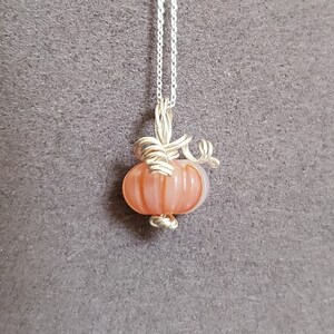 May include: A silver chain necklace with a peach-colored glass pumpkin pendant. The pumpkin has a wire stem and is detailed with lines.