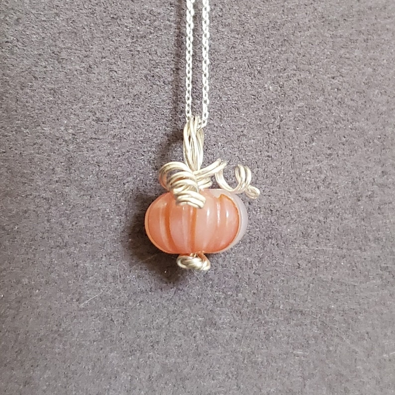 May include: A silver chain necklace with a peach-colored glass pumpkin pendant. The pumpkin has a wire stem and is detailed with lines.