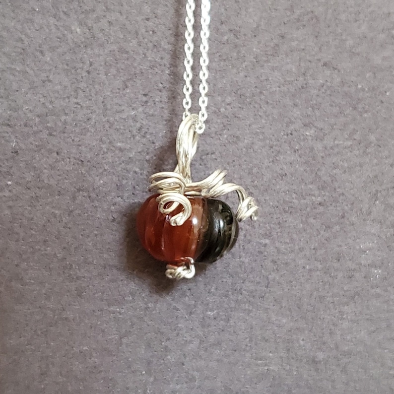 May include: A silver chain necklace with a wire-wrapped pumpkin charm. The pumpkin is made of two glass beads, one brown and one orange.