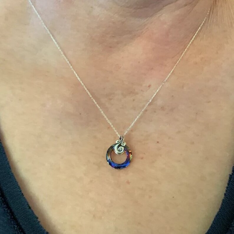 May include: A silver necklace with a pendant featuring a colorful, circular design. The pendant has a swirling, decorative top element and a vibrant mix of blue, red, and orange hues within the circle. The necklace is worn on a person.