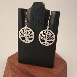 May include: A pair of earrings featuring a tree of life design. The earrings are circular with a detailed cutout pattern, and are made of a pearlescent material. They hang from silver-colored hooks and are displayed on a black stand.