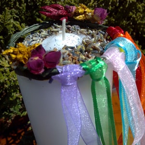 May include: A white candle with a silver glitter top is surrounded by dried flowers and colourful ribbons. The candle is in a clear glass jar.