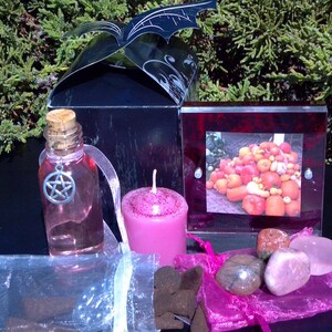 May include: A black gift box with a bat-shaped lid, a pink candle, a clear glass bottle with a pink liquid and a silver pentagram charm, a pink and red picture frame with a photo of pumpkins, and a pink organza bag with pink and brown stones.