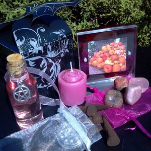 May include: A witchy gift set with a black box, a pink candle, a bottle of pink liquid with a pentagram charm, a small bag of incense, and a frame with a picture of pumpkins.