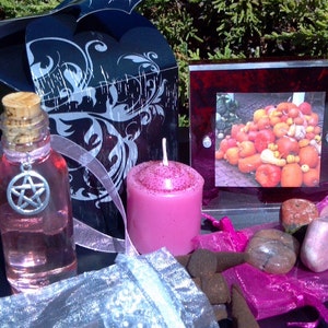 May include: A pink candle, a bottle of pink liquid with a pentagram charm, a small bag of incense cones, a pink and white bag, and a framed picture of a pile of gourds.