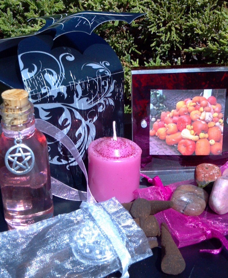 May include: A pink candle, a bottle of pink liquid with a pentagram charm, a small bag of incense cones, a pink and white bag, and a framed picture of a pile of gourds.