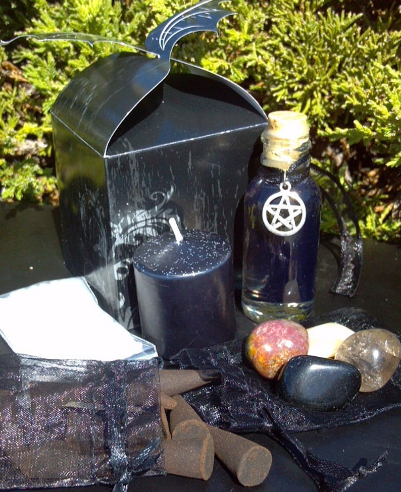May include: A black gift box with bat-wing shaped flaps, a small black pillar candle, a clear glass bottle filled with dark blue liquid and a silver pentacle charm, three polished stones, and a black mesh bag containing brown incense cones.