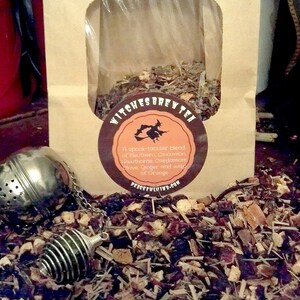 May include: A brown paper bag with a clear window, filled with loose leaf tea. A round label reads "Witches Brew Tea" with a witch graphic. A silver tea infuser is in the foreground, surrounded by tea leaves. The label lists the ingredients.