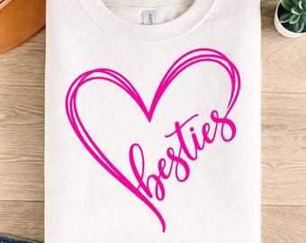 May include: White crewneck sweatshirt with a large pink heart design and the word "besties" in a cursive font. The sweatshirt is folded neatly, with a small plant and a brown leather item visible in the background.
