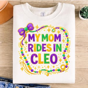 May include: White sweatshirt with the text "MY MOM RIDES IN CLEO" in purple, green, and yellow. The design includes a purple bow, gold stars, and colorful beads, evoking a festive theme.