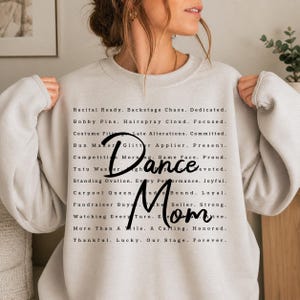 May include: A light beige sweatshirt with the words "Dance Mom" in black script. The sweatshirt has a list of dance-related phrases in a black, sans-serif font.